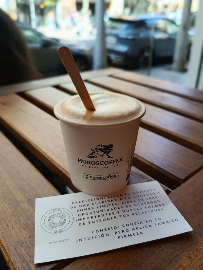Un café con leche de HOROSCOFFEE sobre una mesa de madera, junto a la tarjeta con la predicción del mes y el consejo «confía en tu intuición»